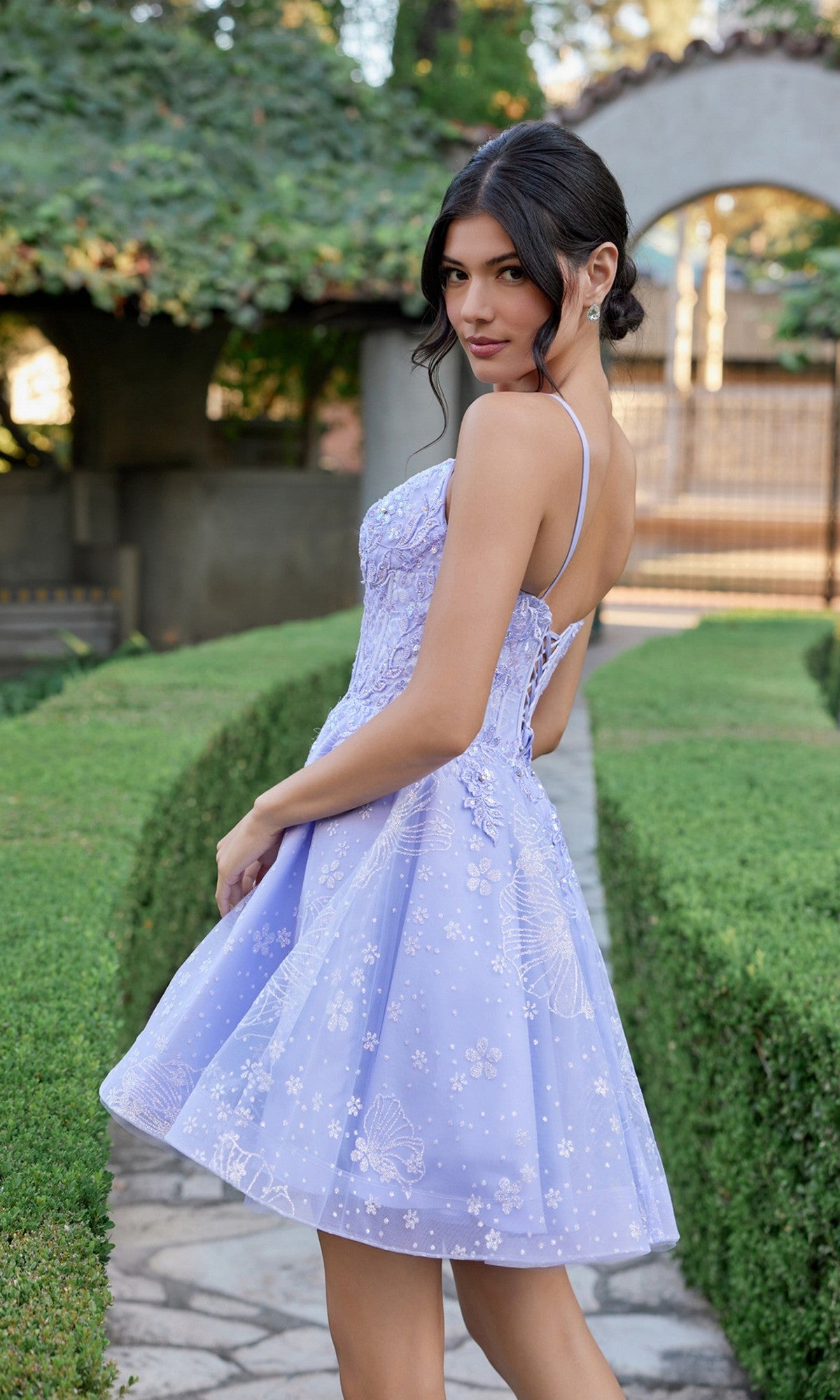 Wearing the Juliet JT985L Short Homecoming Dress—a light purple, sleeveless style with floral patterns and a delicate corset bodice—a woman stands on a garden path, glancing back over her shoulder.